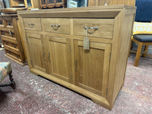 Load image into Gallery viewer, The Bevel Natural Solid Oak Large sideboard is a highly-functional unit with a refreshingly simple design. Made from beautiful solid oak timbers, it is characterised by the bevelled frame with mitred joints and gracefully angled feet. The two doors on the left side of the unit open to reveal a conjoined cupboard space, while the remaining conceals a height-adjustable shelf for taller items. The three large drawers are ideal for storing linen or cutlery and have all been made using traditional dovetail joint