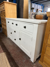 Load image into Gallery viewer, This white-painted oak sideboard combines vintage charm with practical storage. Structurally sound and in good overall condition, it bears gentle signs of use—light marks and scuffs that add character rather than detract from its appeal. Features six drawers with dovetail joins and two cupboards beneath, offering generous storage for living rooms, hallways, or dining spaces. The soft white finish brightens any interior while the solid oak construction ensures lasting durability. A characterful piece that wo