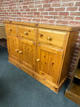 Load image into Gallery viewer, A characterful solid pine sideboard combining vintage charm with practical storage. This well-maintained piece features three dovetail-jointed drawers above single and double cupboards, offering generous space for dining essentials or living room display. Light patina marks reflect its authentic heritage—a testament to quality craftsmanship that's built to last. Perfect for adding warmth and functionality to traditional or eclectic interiors.
Delivery Available
Overall Dimensions
132cm wide x 42cm deep x