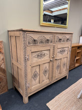 Load image into Gallery viewer, This beautifully carved oak sideboard showcases authentic craftsmanship with dovetail jointed drawers and solid construction. Stripped to reveal natural wood grain, it presents a blank canvas for your finishing touch—a protective wax coat will enhance both appearance and durability. The hinged lift-top design adds practical versatility, while two spacious cupboards and two drawers provide generous storage. A striking focal point for any room, this piece combines period character with functional design.
Del