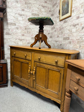 Load image into Gallery viewer, A classic Ercol elm sideboard with solid construction and authentic vintage character. Structurally sound with minor patina marks befitting its age. Features two dovetail-jointed drawers above a generous double cupboard—ideal for dining room storage or hallway display. Timeless design and quality craftsmanship make this a versatile addition to period or contemporary interiors.
Delivery Available
Overall Dimensions
123cm wide x 46cm deep x 84cm high 