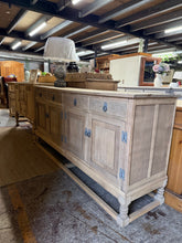 Load image into Gallery viewer, Authentic solid oak sideboard, stripped to natural wood and ready for your finishing touches. This preloved piece showcases genuine character—subtle wear marks and a slight dip to the top that adds to its rustic appeal. Four dovetail-jointed drawers sit above two double cupboards, offering generous storage for dining rooms or living spaces. A light wax treatment will enhance the grain and protect the surface. Perfect for those who appreciate vintage craftsmanship and unique furnishings with personality.
De