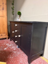 Load image into Gallery viewer, A substantial black sideboard offering authentic character and practical storage. Built solid and fully functional, this piece shows honest wear befitting a pre-loved furniture find. Features include two cupboards and four small drawers, providing generous organisation space. One leg is unpainted so could benefit from a coat of black paint to match.
Delivery Available
Overall Dimensions
139cm wide x 51cm deep x 86cm high