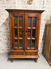Load image into Gallery viewer, This charming hardwood display cupboard combines practical storage with vintage character. Structurally sound with two internal shelves and two short drawers, it offers ample space for displaying treasures or storing tableware. The glass doors feature delicate hand-painted floral details added by previous owners, creating a unique focal point. Minor surface scratches and a missing corner piece are present but this can be reattached. Perfect for those seeking distinctive vintage furniture with genuine patina