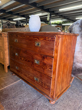 Load image into Gallery viewer, This characterful pitch pine campaign-style chest of drawers combines solid construction with authentic vintage appeal. Featuring three spacious dovetail-jointed drawers, it offers practical storage with period charm. The piece shows its age beautifully through natural patina and marks that enhance its character. Please note: the top drawer requires gentle lifting to close smoothly—a minor quirk that speaks to its genuine heritage and craftsmanship.
Delivery Available
Overall Dimensions
126cm wide x 50cm