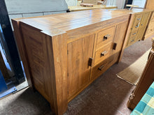 Load image into Gallery viewer, This beautiful, solid mango wood sideboard is sturdy and in good overall condition. While a bit of the veneer on the top is peeling, it doesn’t impact its functionality. It features three short drawers with dovetail joints and two single-door cupboards, each with one shelf, providing ample storage space.
Overall Dimensions
149cm wide x 60cm deep x 85cm high
DELIVERY AVAILABLE
For our other items see our website https://fossewayfurniture.co.uk
collection from our shop at cv36, our collection zone