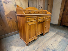 Load image into Gallery viewer, A characterful solid pine sideboard with authentic vintage appeal. Structurally sound and in good overall condition, this piece shows honest wear consistent with its age—minor marks add to its charm rather than detract from it. Generously proportioned storage includes six small drawers, two longer dovetail-jointed drawers, and three single cupboards, offering flexible organisation for any room. The removable upper section sits independently, allowing you to adapt the layout to suit your space.
Delive