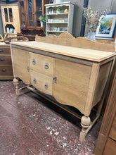 Load image into Gallery viewer, Stripped back to bare oak, this sideboard displays natural wood tones and authentic grain patterns. Two cupboards and two dovetail-jointed drawers provide ample storage capacity. The piece carries expected signs of age with minor surface marks that enhance its vintage character. Optional wax finish would offer additional protection. Supplied with key. Complements country, farmhouse, and traditional home décor schemes beautifully.

Delivery Available

Overall Dimensions

137cm wide x 48cm deep x 92cm / 109cm
