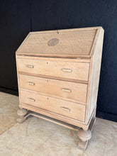 Load image into Gallery viewer, This beautiful oak bureau showcases authentic craftsmanship with its natural finish and period character. The stripped-back wood reveals the grain and patina of quality construction, though a protective wax coat is recommended to maintain its appearance. Practical storage is provided by three generously proportioned dovetail-jointed drawers, whilst the pull-down writing desk offers a functional workspace. A decorative plaque adorns the top, adding historical charm to this statement piece. Ideal for those se
