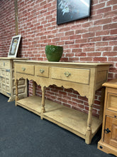 Load image into Gallery viewer, A charming stripped oak hall table with authentic character. This piece has been carefully stripped back to reveal the natural wood grain, creating a fresh, versatile foundation for your décor. Three dovetail-jointed drawers—two long and one short—provide practical storage, while a lower shelf offers additional space for baskets or display. Structurally sound and in good condition, it would benefit from a protective coat of wax to enhance and preserve the wood. Perfect for entryways, living rooms, or as a s
