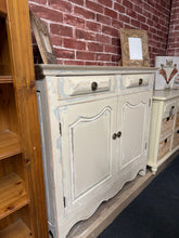 Load image into Gallery viewer, A charming cream distressed sideboard that combines vintage character with practical storage. Structurally sound and in good overall condition, this narrow piece features two drawers positioned above a double cupboard with internal shelf—perfect for concealing everyday essentials while keeping them within easy reach. The gentle wear and patina add authentic period appeal, making it an ideal accent for farmhouse, shabby chic, or eclectic interiors. A versatile addition to hallways, living rooms, or dining sp
