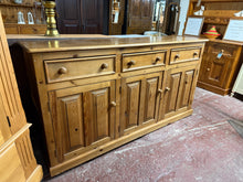 Load image into Gallery viewer, This substantial solid pine sideboard combines practical storage with authentic character. Featuring three dovetail-jointed drawers positioned above a single and double cupboard with internal shelving, it offers versatile space for dining essentials. The piece is structurally sound and fully functional. As a pre-loved item, it displays the natural patina of age with minor surface marks that enhance its vintage appeal—these can be easily refreshed with furniture wax if desired. A genuine, hardwearing additio
