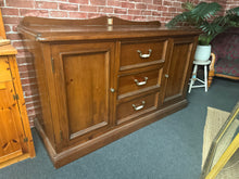 Load image into Gallery viewer, A charming solid pine sideboard that combines practicality with character. This well-built piece features three dovetail-jointed drawers perfect for everyday items, plus two cupboards with shelves for flexible storage solutions. The natural patina and minor marks add authentic vintage appeal, while the sturdy construction ensures reliable everyday use. An excellent addition to any home seeking functional, timeless furniture.

Overall Dimensions

160cm wide x 57cm deep x 91cm high to top of unit

DELIVERY AV
