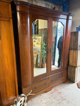 Load image into Gallery viewer, This substantial mahogany wardrobe combines classic design with practical functionality. The piece features two bevelled-edge mirrors across the front, creating an elegant focal point. Inside, you'll find two single cupboards, each equipped with a hanging rail and multiple hooks for organised storage, plus two dovetail-jointed drawers at the base for additional space. The wardrobe shows its age with minor surface marks, but remains structurally robust and fully functional. For convenience, it disassembles i
