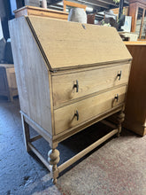 Load image into Gallery viewer, This stripped oak bureau reveals beautiful natural wood grain throughout. The piece features a pull-down writing desk with two spacious dovetail-jointed drawers beneath, offering practical storage and workspace. Recently stripped to bare wood, it's ready for finishing with wax or your preferred protective coating. Please note: some veneer shows minor peeling, which doesn't impact functionality. A characterful piece with solid construction, ideal for those who appreciate authentic vintage furniture with char
