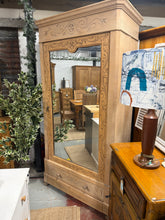 Load image into Gallery viewer, This striking oak armoire makes a bold focal point with its generous proportions and exposed wood grain. Currently stripped and awaiting your preferred wax finish for lasting protection, it retains authentic patina that speaks to its age and use. Internally, a hanging rail accommodates clothing whilst a dovetail-jointed drawer provides additional storage at the base. Surface marks and a minor imperfection on the mirror reflect its genuine vintage character rather than detract from its appeal. Practical desi
