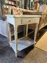 Load image into Gallery viewer, A practical white side table with genuine character. This piece is structurally solid and fully functional, featuring one drawer and a lower shelf—ideal for keeping essentials within reach. As a pre-loved item, it bears the honest marks of use, though nothing detracts from its everyday appeal.&nbsp;

Delivery Available

Overall Dimensions

60cm wide x 41cm deep x 76cm high

