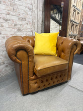Load image into Gallery viewer, A classic Chesterfield club chair in warm tan leather, offering timeless style and comfort. The frame is structurally sound with minor surface marks that can be easily minimised with a leather wax treatment. The deep button-tufted back and rolled arms provide authentic period charm, making it an ideal centrepiece for a gentleman's study, library, or traditional living room. Note: yellow cushion shown is not included. Ideal for collectors of vintage furniture or those seeking a statement piece with character
