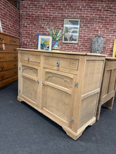 Load image into Gallery viewer, A characterful stripped oak sideboard with genuine vintage appeal. Features two dovetail jointed drawers and a double cupboard for substantial storage. The natural wood finish may benefit from wax treatment to enhance protection and patina. Structurally sound and in good condition. Please note: one back leg is missing and will require a support or replacement before use. Ideal for those seeking authentic period furniture with character and functionality.

Delivery Available

Overall Dimensions

124cm wide x
