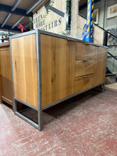 Load image into Gallery viewer, This industrial-style oak sideboard combines authentic character with practical storage. Structurally sound and well-maintained, it features two single cupboards each with a shelf and three dovetail-jointed drawers—ideal for displaying tableware or storing linens. Light wear is present but adds to its rustic charm. A versatile piece that complements country, farmhouse, and eclectic interiors with understated elegance. Originally from Oak Furniture Land.

Delivery Available

Overall Dimensions

160cm wide x 
