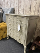 Load image into Gallery viewer, This elegant French-style chest of drawers features a distinctive serpentine front and soft, distressed light grey finish that adds timeless character to any bedroom. Structurally sound and well-maintained, it showcases traditional craftsmanship with three deep dovetail-jointed drawers. The top two drawers include fixed dividers, perfect for organising jewellery, accessories and smaller items with ease. Minor patina marks enhance its authentic vintage appeal. A versatile statement piece that combines classi

