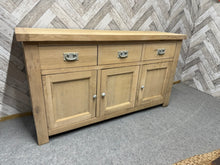 Load image into Gallery viewer, A solid oak sideboard stripped to bare wood, revealing beautiful natural grain throughout. Features three dovetail-jointed drawers plus a double and single cupboard, offering substantial storage capacity. The piece is ready for finishing—a protective wax treatment is advisable to preserve the wood. The back panel has been modified by previous owners and would benefit from repair or replacement with plywood.

Delivery Available

Overall Dimensions

138cm wide x 43cm deep x 77cm high

